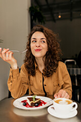 Charming excited European girl with curls sitting in cafeteria and enjoying dessert and coffee....