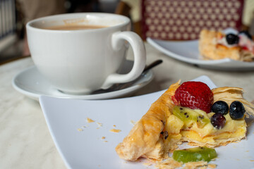 A white porcelain cup and saucer with a small tea spoon on the saucer. The mug is filled with hot coffee and cream. There are square plates on the white marble dining table with fruit and pastry.