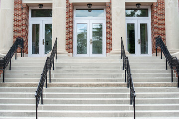 Concrete steps lead to a red double door of a historic building. The wall of the building is made of a light grey granite block. There are four metal handrails dividing the stairs to the entrance.