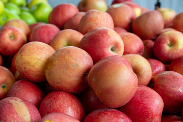A stack or bulk supply of large red ripe Fuji apples on a table for sale at a farmer's market. There are green granny smith apples in the background. The red organic apples are fresh sweet produce.