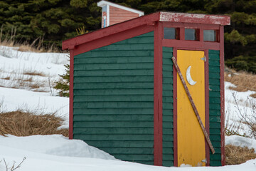 A small yellow and green building with a wooden door. The outhouse or outdoor toilet is in a meadow...