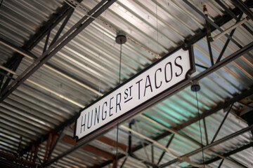 A white with black lettering sign hanging from the ceiling of a market. The sign spells hunger street tacos. The interior of the industrial warehouse ceiling is silver metal with a black grid system.