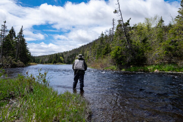 A male salmon angler stands in a river up to his waist with a long fishing rod wearing waders, a fly hat, and a vest. The background is trees and a riverbank. The blue water has a small ripple on top.