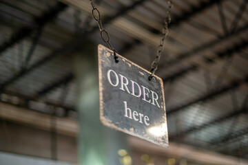An order here sign made of metal with white letters on a black background hung over the counter and cash register. The directional sign hangs from a metal interior roof of a restaurant. 