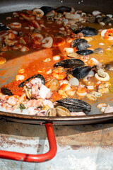 A large and wide carbon steel garcima paella pan filled with seafood; mussels, clams, squid, scallops, and shrimp cooking on a stove. The seafood is stewing in a red tomato broth prior to adding rice.