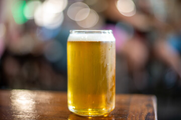 A clear drinking glass in the shape of a beer can filled with cold froth lager ale. The Belgian pint sits on the edge of a wooden patio table at a microbrewery. The background has multicolored lights. - Powered by Adobe
