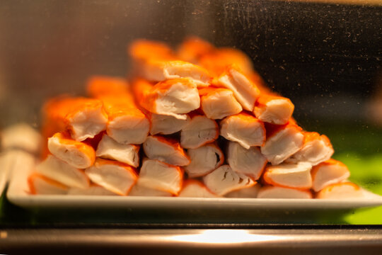 Crab Or Krab Sticks Are Stacked In A Cooler. The Red And White Colored Imitation Seafood Crab Legs Are Made From Japanese Spider Crab Protein Meat.  The Stack Of Legs Is On A White Plate.