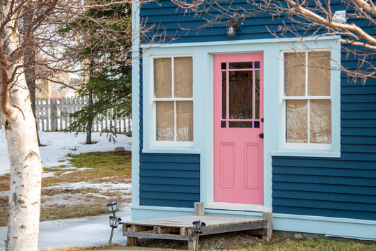 A vintage pink door with a half glass window. The old door has a brass doorknob. There are two glass windows on both sides of the doorway. The yard near the building has grass, snow, and trees. - Powered by Adobe