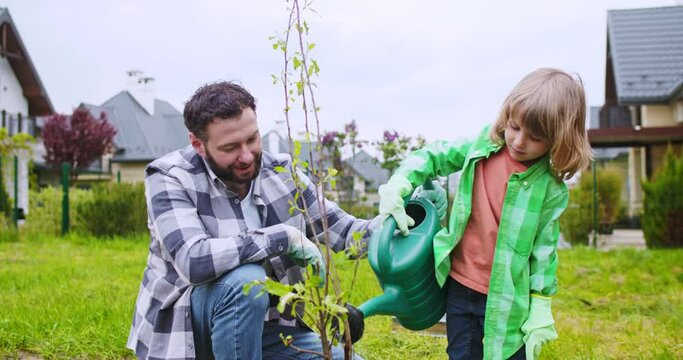 Caucasian father teaching little cute son watering planted small tree at summer house. Kid with dad planting trees and water with pot. Summer work in garden.