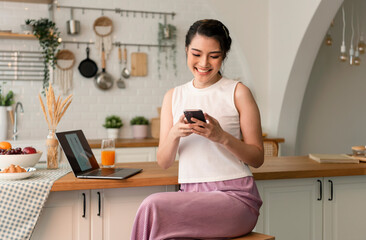 Smiling young asian woman using mobile phone while sitting in kitchen room at home with laptop...