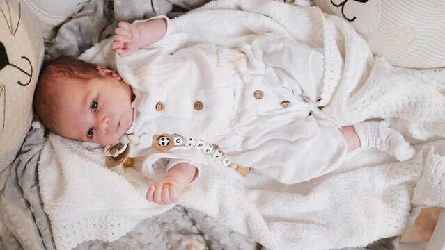 Active newborn baby lies in crib and moves and looks around and explores world. Paternity. Happiness of motherhood. Child is in romper and covered with blanket. Pacifier nearby. Care and concern