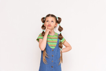 A little girl looks up thoughtfully with funny ponytails and a summer denim sundress on a white isolated background,