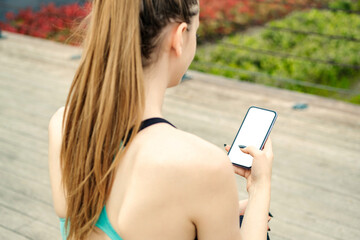 Brunette sportive woman wearing black sports bra standing on city park, outdoors hands holding phone touching finger mockup white blank display, mobile application tech concept, over shoulder view.