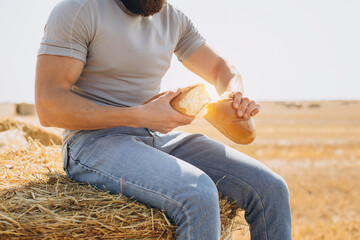 Bearded man farmer in jeans holding fresh fragrant bread tears it in half sitting on top of a bale...