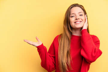 Young caucasian woman isolated on yellow background holds copy space on a palm, keep hand over cheek. Amazed and delighted.