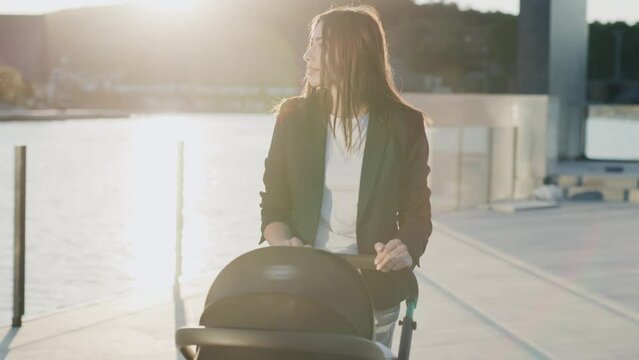 Cute happy woman stand in the sunny park with a stroller near lake. Looking around smiling. Motherhood. Close up. Slow motion