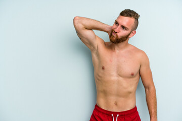 Young caucasian man wearing a swimsuit isolated on blue background touching back of head, thinking and making a choice.