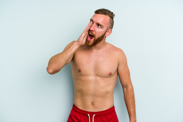 Young caucasian man wearing a swimsuit isolated on blue background shouting and holding palm near opened mouth.