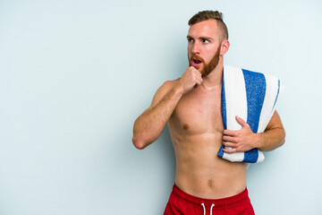 Young caucasian man going to the beach holding a towel isolated on blue background looking sideways with doubtful and skeptical expression.