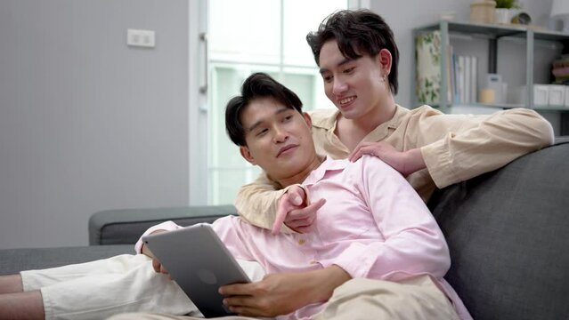 Cute Male Gay Couple Spend Time at Home. Young happy lgbtq couple Lying Down on a Sofa and Use a tablet, doing online shopping in ecommerce app together, having fun checking social media together