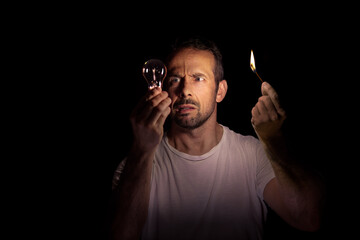 Worried man looking at a light bulb with a match in the dark. Concept of blackout. Selective focus.