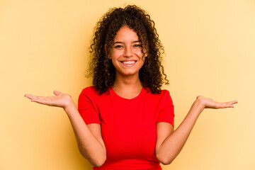 Young Brazilian woman isolated on yellow background makes scale with arms, feels happy and...