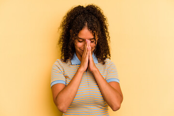 Young Brazilian woman isolated on yellow background praying, showing devotion, religious person looking for divine inspiration.