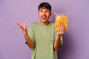 Young hispanic man holding crisps isolated on purple background receiving a pleasant surprise,...