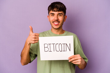 Young hispanic man holding bitcoin placard isolated on purple background smiling and raising thumb...
