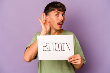 Young hispanic man holding bitcoin placard isolated on purple background trying to listening a gossip.