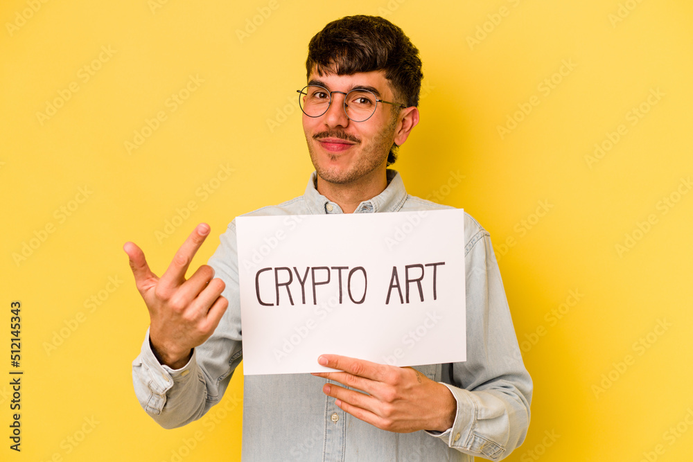 Wall mural Young hispanic man holding crypto art placard isolated on yellow background pointing with finger at you as if inviting come closer.