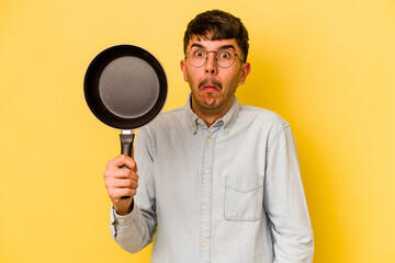 Young hispanic cooker holding frying pan isolated on yellow background shrugs shoulders and open...