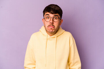 Young caucasian man isolated on purple background shrugs shoulders and open eyes confused.