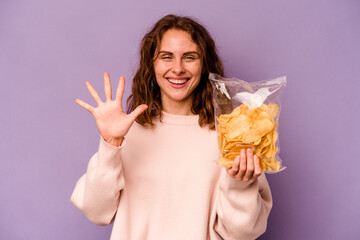 Young caucasian woman holding a bag of chips isolated on purple background smiling cheerful showing number five with fingers.