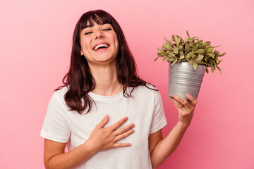 Young caucasian woman holding a plant isolated on pink background laughs out loudly keeping hand on...
