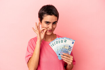 Young caucasian woman holding banknotes isolated on pink background with fingers on lips keeping a secret.