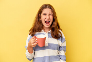 Little caucasian girl holding a pink mug isolated on yellow background screaming very angry and...