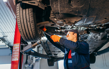 Young car mechanic at repair service station inspecting car wheel and suspension detail of lifted automobile. Bottom view.