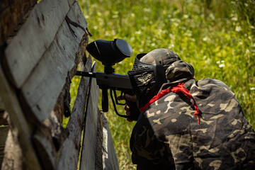 a paintball player sits behind a wooden shelter in the summer in the grass and shoots, visas from behind. a game of paintball, a man in a helmet and glasses holds a gun. close-up