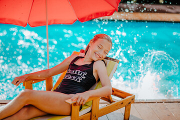 Girl posing on beach chair with red umbrella against splashing pool. Sunny day