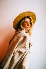 Portrait of attractive caucasian woman in eyeglasses and wicker hat wearing in stylish outfit, posing on white wall background.