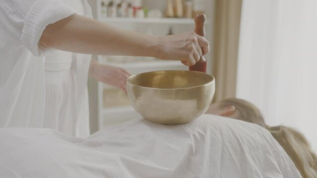 Masseuse twists stick around singing bowl during sound therapy session spa.