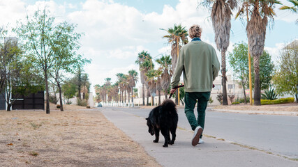 Man in green sweater walks his big black dog down the street on a leash