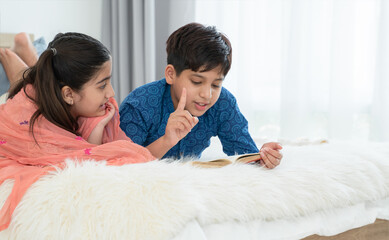 Two happy Indian brother and sister in traditional clothing lying on bed and reading book, having fun together at home. Education, Siblings relationship concept