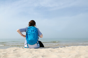 Dziewczyna, kobieta z plecakiem siedzi na piaszczystej morskiej plaży. Wpatruje się w horyzont. Lato, pastelowe kolory, przydymione błękitne niebo - obrazy, fototapety, plakaty