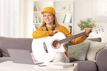 Young female sitting on a sofa at home with an acoustic guitar and a laptop computer