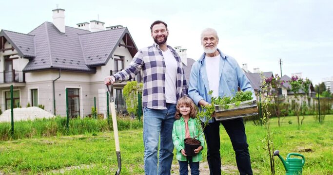 Portrait of cute happy small boy with plant in hands smiling to camera and standing together with father and grandfather. Planting trees and plants in garden at house. Outdoor.