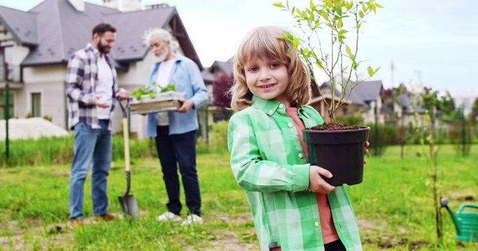 Portrait of cute Caucasian happy small boy with plant in pot in hands smiling to camera. Father and grandfather talking on background. Planting trees and plants in garden at house. Outdoors.