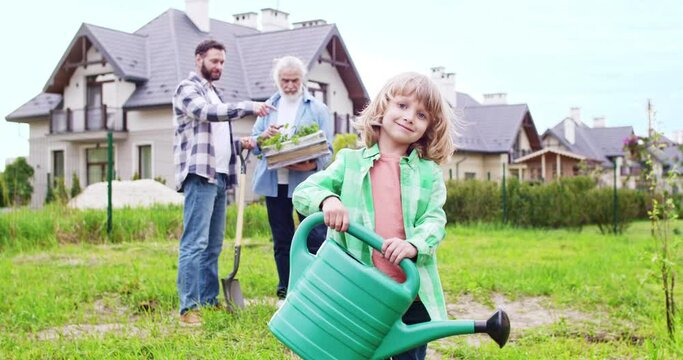 Portrait shot of cute Caucasian cheerful small boy with watering pot in hands smiling to camera. Father and grandfather talking on background. Planting trees and plants in garden at country house.