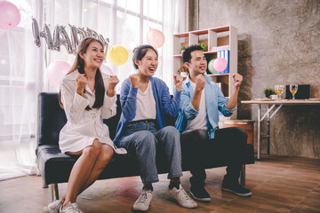 A group of Asian friends sit at home watching on the sofa and cheering for a sporting match on TV together. Excited male and female sports fans celebrate team sports victories at national games.